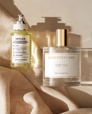 Dwa flakony perfum: REPLICA Maison Margiela z żółtą cieczą i ZARKOPERFUME Oud’ish eau de parfum, oba stojące na beżowej tkaninie.