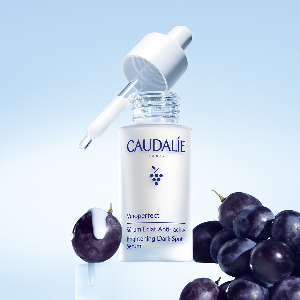 Biała butelka serum Caudalie Vinoperfect Brightening Dark Spot Serum z pipetą na tle niebieskiego gradientu, otoczona ciemnymi winogronami.