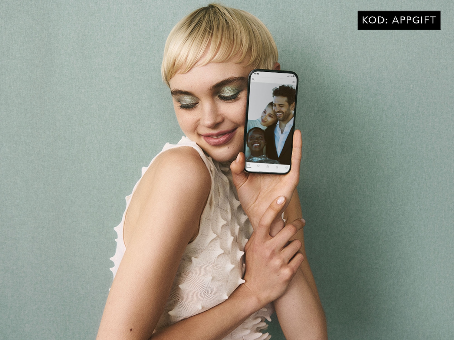 Kobieta z krótkimi blond włosami trzyma telefon, na ekranie którego widać uśmiechnięte twarze; kod rabatowy: APPGIFT. Prezent niespodzianka za pierwsze zakupy w aplikacji o wartości min. 49 zł.