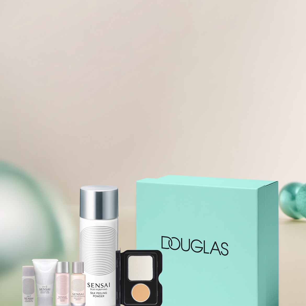Zestaw kosmetyków Sensai, w tym peeling w pudrze w białej butelce, kremy w tubkach i puder w czarnym opakowaniu, obok pudełko Douglas w kolorze morskim.