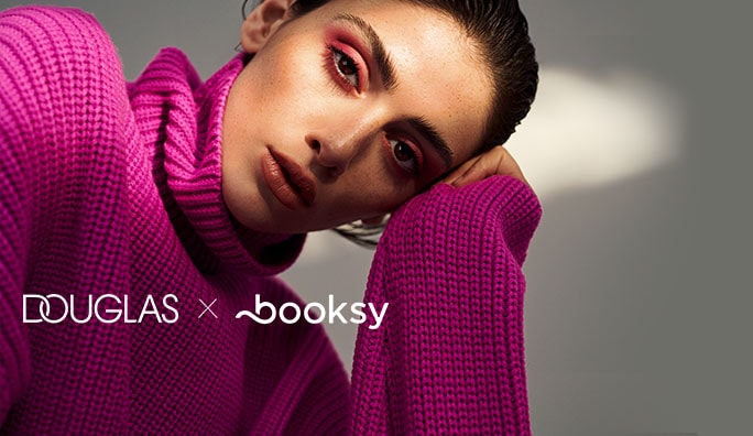 Na zdjęciu kobieta w różowym swetrze z golfem, z makijażem w odcieniach różu, prezentuje się na tle z logo Douglas x booksy.