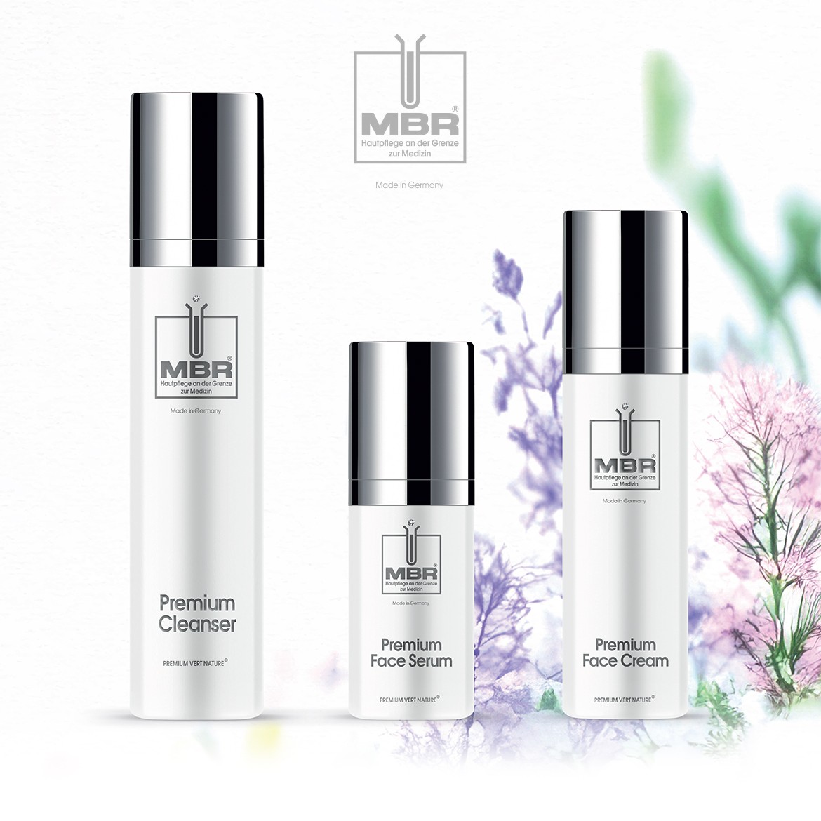 Trzy białe butelki produktów do pielęgnacji skóry MBR Premium Vert Nature: Cleanser, Face Serum i Face Cream, z srebrnymi nakrętkami, na tle kwiatów.