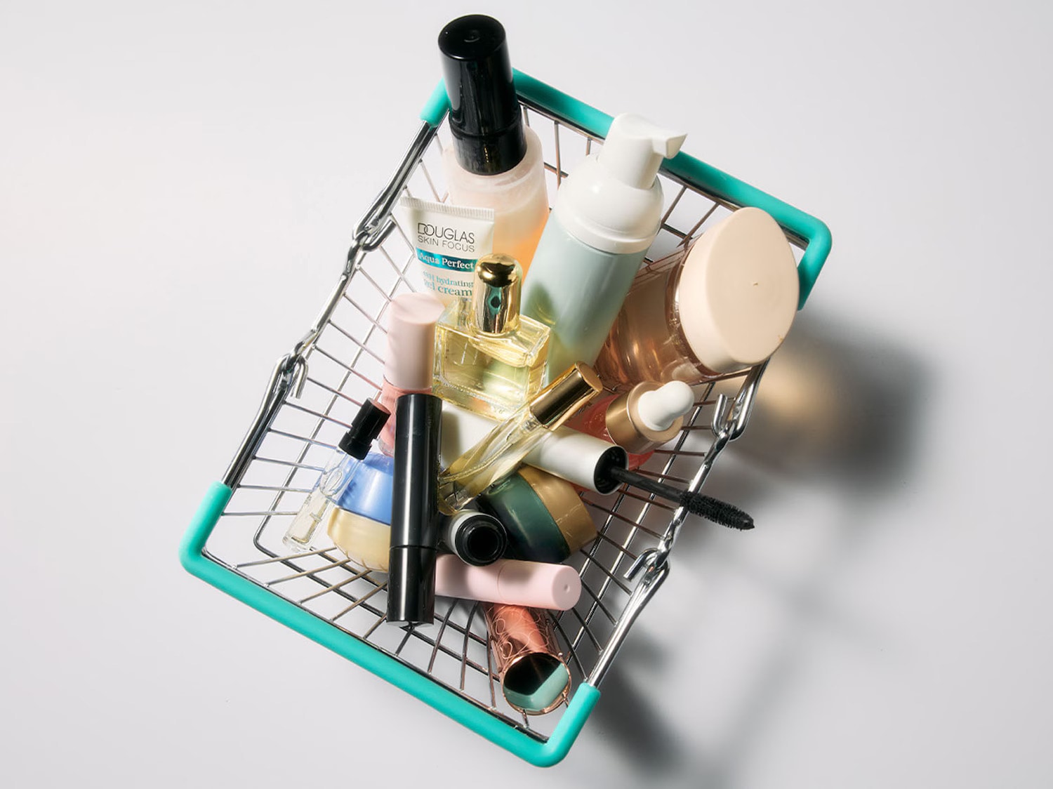 Koszyk na zakupy wypełniony różnorodnymi produktami do makijażu i pielęgnacji skóry, w tym kremem nawilżającym Douglas Skin Focus Aqua Perfect, perfumami, tuszem do rzęs i innymi kosmetykami.