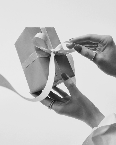Dłonie z czerwonymi paznokciami rozwiązują białą wstążkę na szarym pudełku prezentowym, ozdobione pierścionkami.