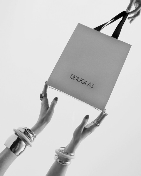 Elegancki prezent od Douglas: srebrna torba z czarnymi uchwytami, trzymana przez dłonie ozdobione srebrnymi bransoletkami i pierścionkami, na białym tle.