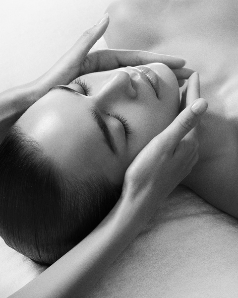 Na czarno-białym zdjęciu widać kobietę z zamkniętymi oczami, której twarz jest delikatnie masowana dłońmi, co sugeruje relaksacyjny zabieg pielęgnacyjny.