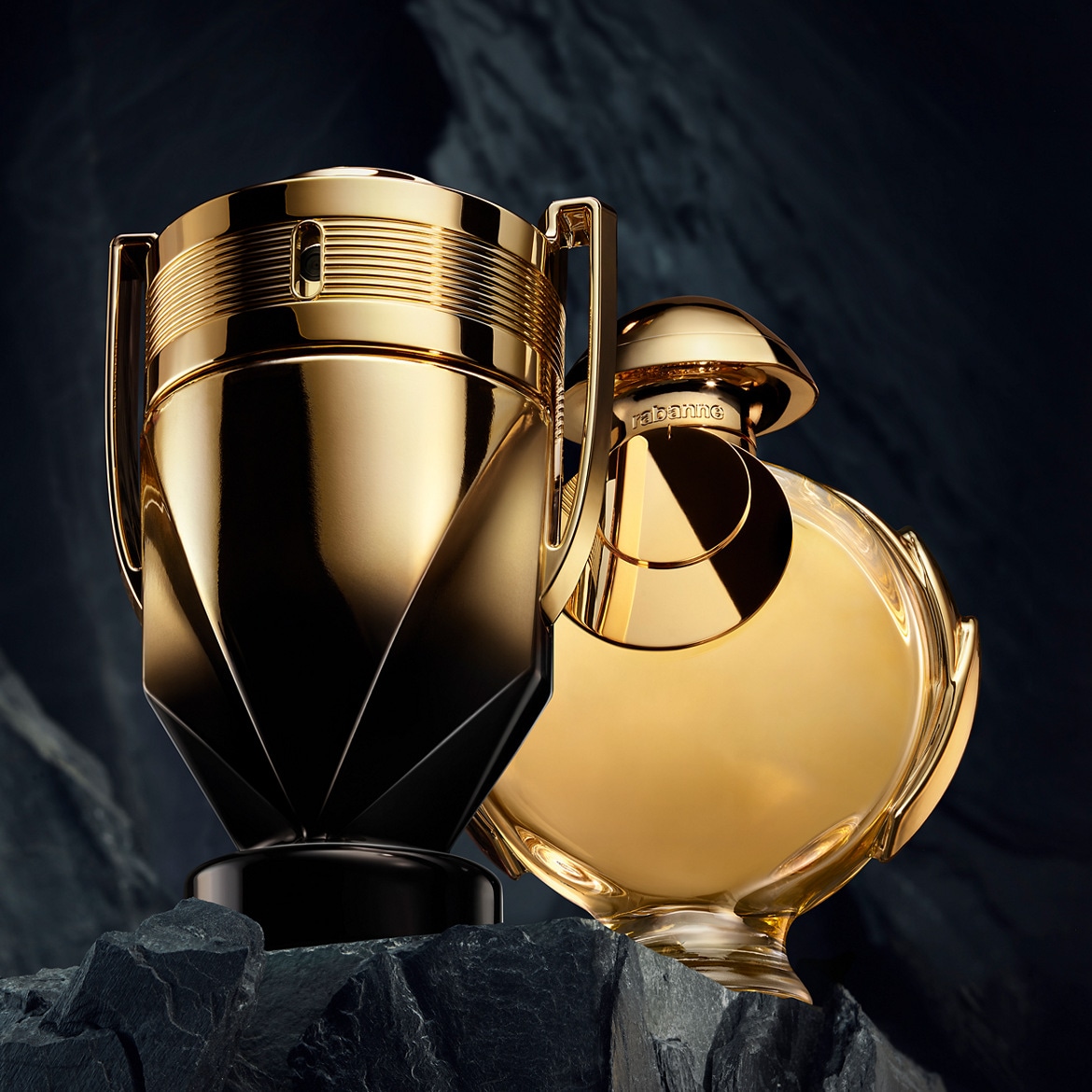 Dwa złote flakony perfum Paco Rabanne, jeden w kształcie pucharu, drugi okrągły, stoją na ciemnych kamieniach na tle ciemnego tła.