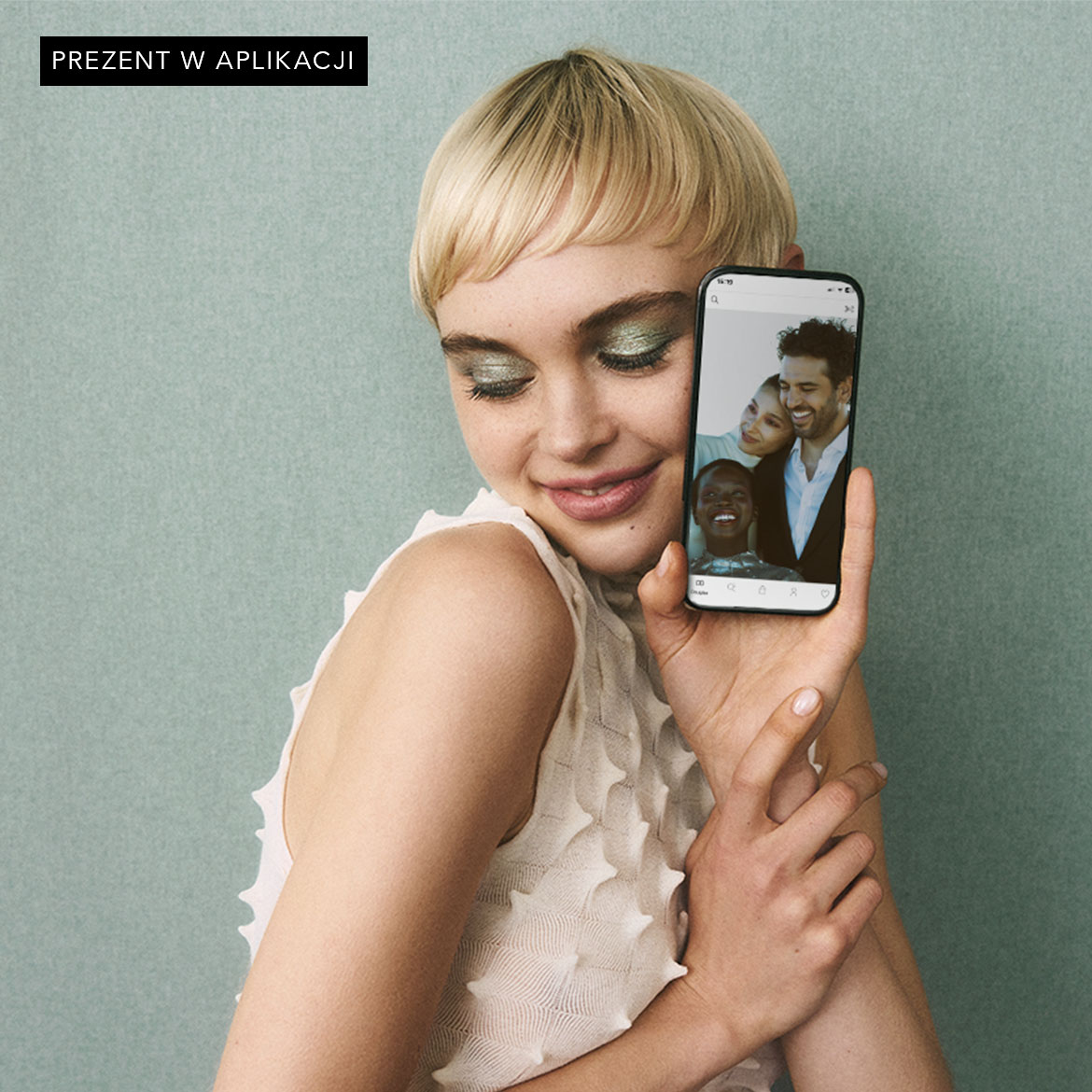 Kobieta z krótkimi blond włosami i zielonym makijażem oczu trzyma telefon, na którego ekranie widać zdjęcia uśmiechniętych osób; napis: Prezent w aplikacji.