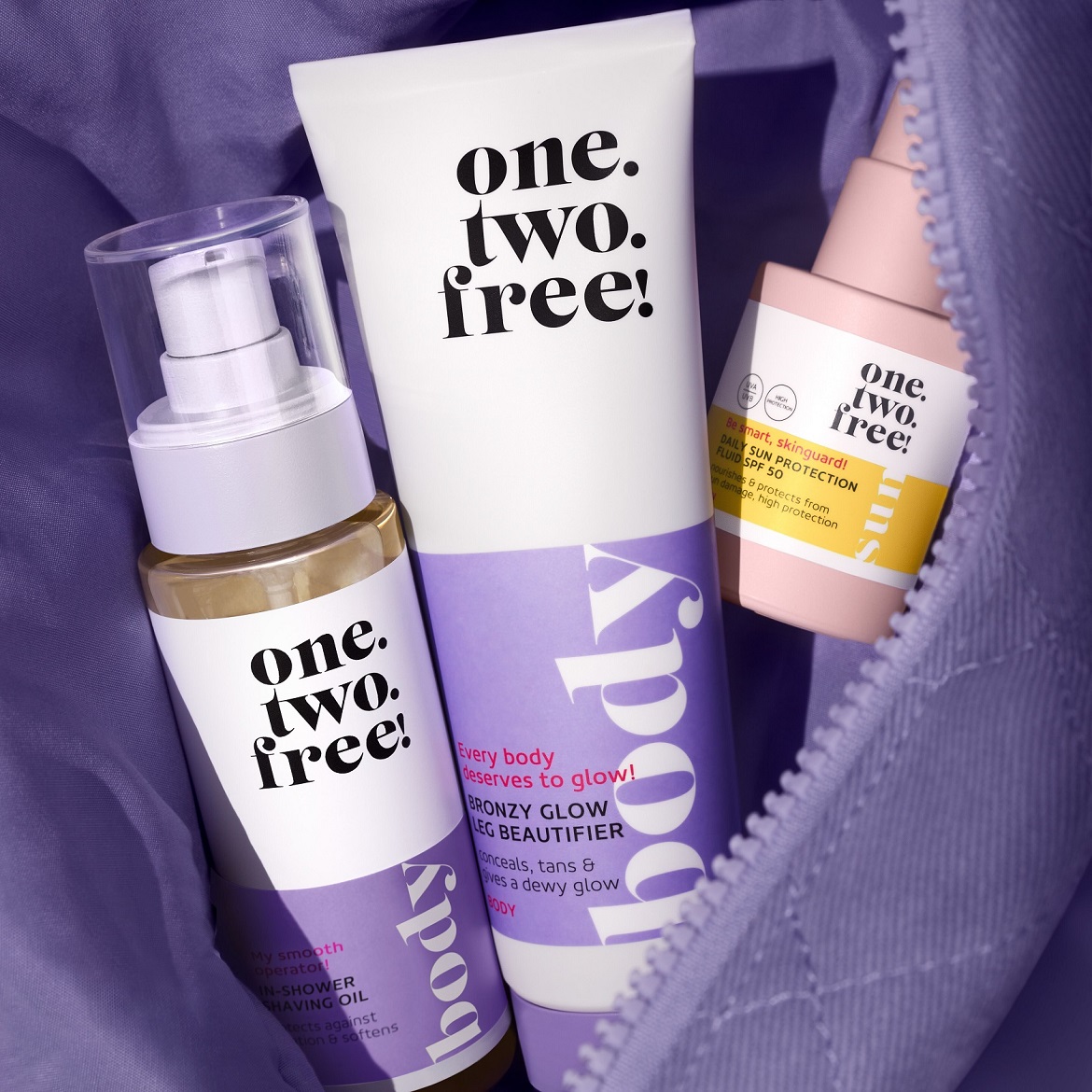 Zestaw kosmetyków One. Two. Free! w fioletowej kosmetyczce obejmuje olejek do golenia, balsam brązujący do nóg i fluid do opalania z filtrem SPF 50, zapewniając kompleksową pielęgnację i ochronę skóry.